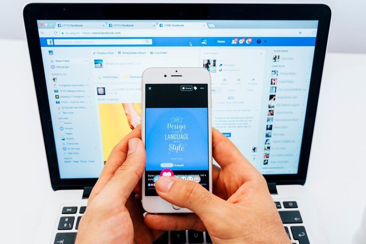 A laptop displaying a Facebook page with two hands holding a phone, interacting with a post. This illustrates the common mistake of using the Boost Post option in Meta Ads.